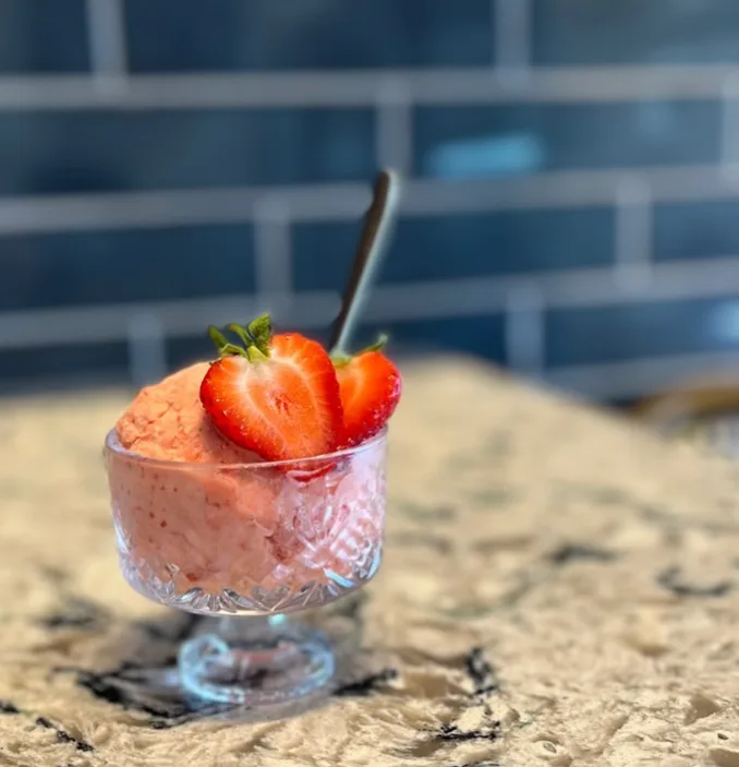 A scoop of strawberry gelato in a glass dessert cup topped with fresh strawberries